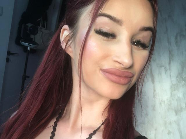 FoxxyAmber with red hair, winking, and pouty lips, wearing a black necklace, has one eyelid closed, long eyelashes, and a subtle pink blush on her fair skin.