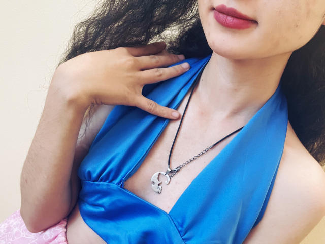 Millinia with curly black hair, wearing a blue halter top, showing her small tits, has pink lipstick, and a silver necklace with a key pendant, touching her chest.