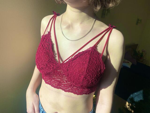 Steisii wearing a red lace bralette with strappy details, showing her small tits and slim, sunlit torso, with a delicate chain necklace and short brown hair.