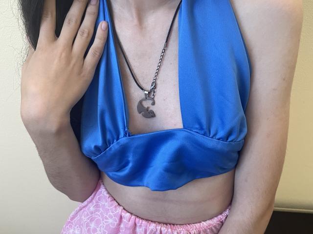 Millinia's blue satin halter top, showing her tits, with a black necklace and pink patterned pants, her hand near her face, light skin, long dark hair.