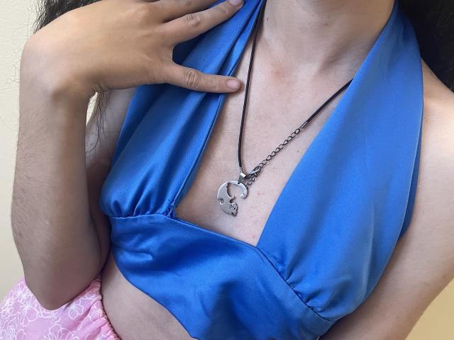Millinia's blue satin halter top reveals her small tits, with a silver necklace hanging between them. Her hand gently touches the top. She's wearing pink patterned pants.