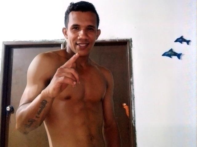 DreamGuy69 sin camisa, con un cuerpo tonificado y musculoso, señalando su boca con un dedo, sonriendo, tiene el pelo corto y negro y un tatuaje en el brazo izquierdo..
