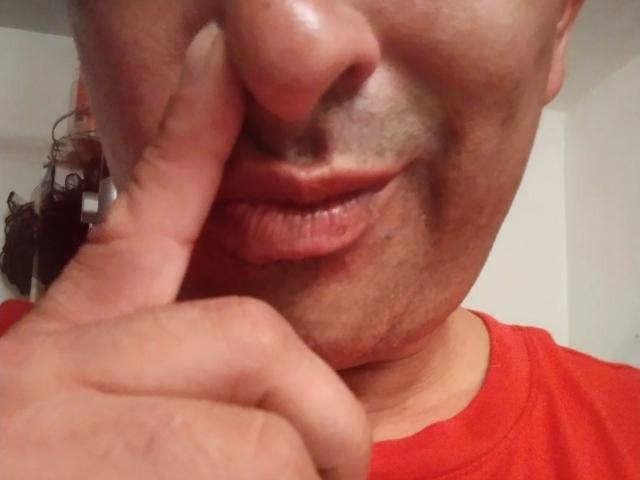 Il viso di Leaveitinsideme-hot, con un dito vicino al naso, indossa una maglietta rossa, mostrando labbra leggermente gonfie e rosse, e un piccolo pizzetto. La sua pelle è leggermente lucida..