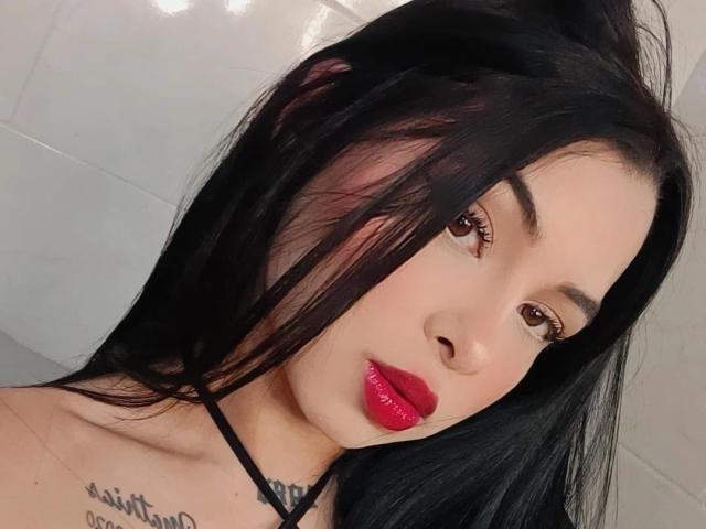 MistressAsshley has black hair, red lipstick, and brown eyes. She's wearing a black halter top, showing a tattoo on her shoulder and a sexy, confident face.