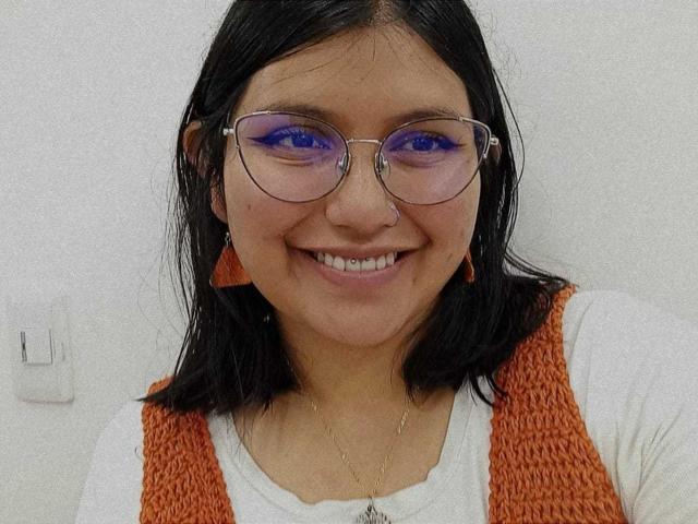 Sara, met zwart haar, paarse oogschaduw, een bril, een oranje gebreide vest, een wit shirt, lachend, een neusring, oorbellen en een ketting, voor een effen witte muur..