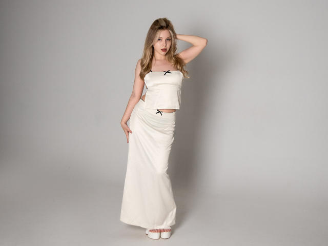DellaLovely with blonde hair, wearing a white strapless top and matching white skirt, standing with one hand on her head, showing her tits and small black bow.