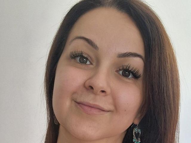 LoraHazel's face with brown hair, light skin, and long eyelashes, smiling slightly. She has small tits and is wearing black earrings. Her brown eyes are looking at the camera.