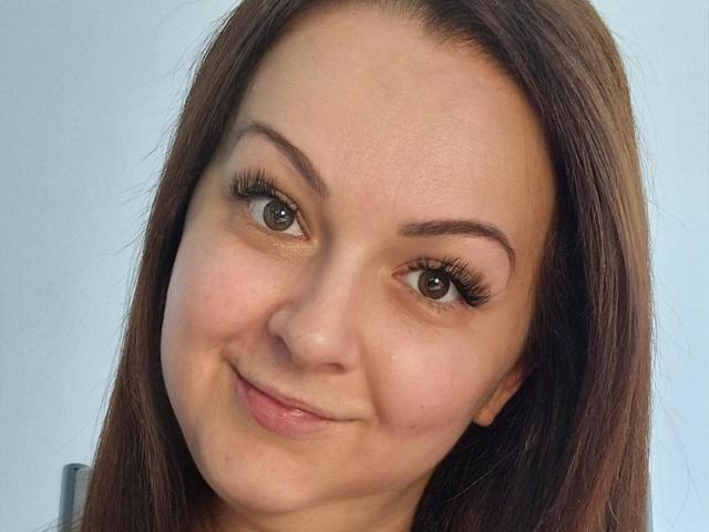 LoraHazel has brown eyes, long brown eyelashes, and a cute smile with slightly flushed cheeks. Her brown hair is straight and she's looking at the camera.