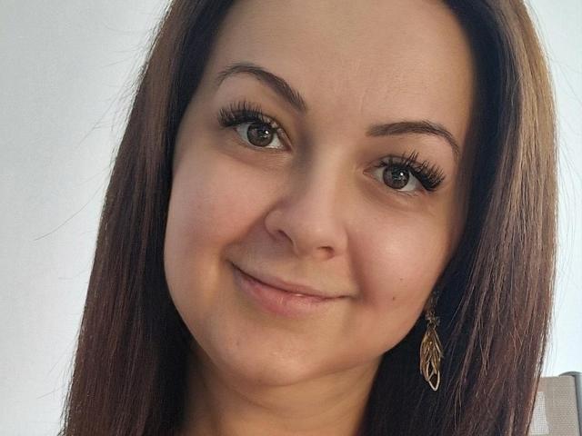 LoraHazel has brown hair, green eyes, and wears a gold leaf earring. She's smiling with long eyelashes and a slight pink blush on her fair skin.