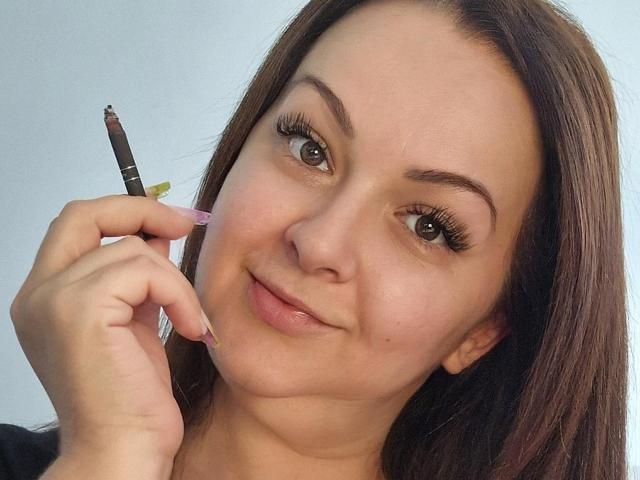 LoraHazel with brown hair, holding a pink-tipped eyeliner, applying it to her right eyelid. She has long eyelashes, a subtle smile, and fair skin.