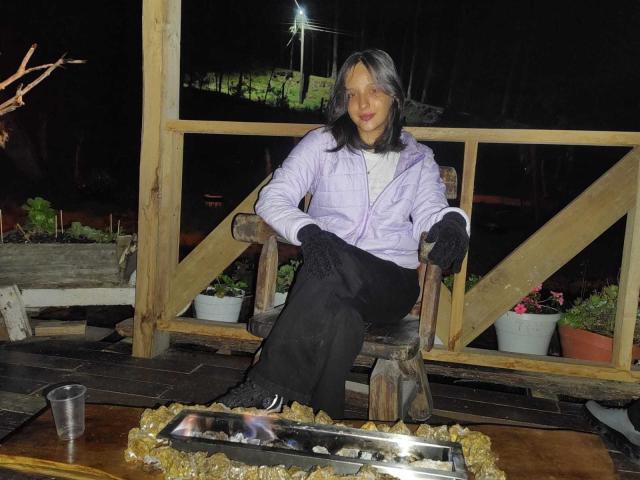 LanaKlaire está sentada em uma cadeira de madeira, vestindo uma jaqueta roxa, calças pretas e luvas, com cabelo grisalho, ao lado de uma fogueira, segurando uma bebida. Noite..