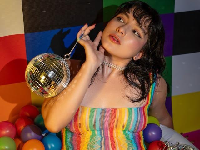 HairyValeri with wet black hair, wearing a rainbow-striped dress, holding a mirror ball, and a pearl necklace, surrounded by colorful balloons, looking up with a sultry expression.