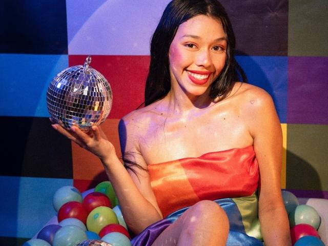 RoseSexAndFun avec de belles seins, portant une robe rouge-orange en satin sans bretelles, tenant une boule de miroir argentée, assise dans un bac à balles coloré, souriant avec du rouge à lèvres rouge..