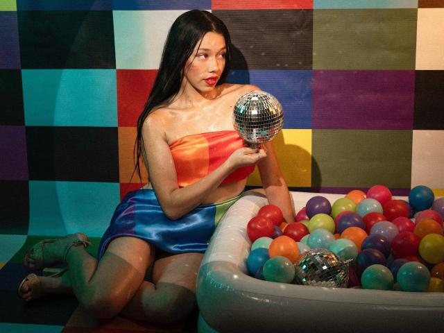 RoseSexAndFun, avec de longs cheveux noirs, une robe brillante orange et bleue, tenant une boule de miroir argentée, agenouillée dans une piscine gonflable colorée remplie de balles multicolores..