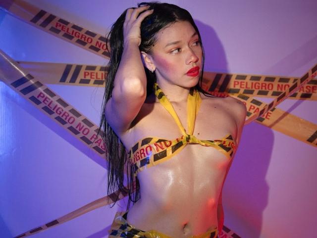 RoseSexAndFun with wet black hair, red lipstick, and yellow "caution" tape bikini, holding hair, showing shiny, glistening, sweaty, bare tits and small, hard nipples.