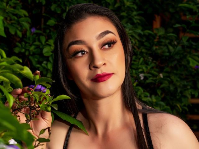 EmmaLovesX with dark hair, red lipstick, and brown eyes, holding purple flowers, wearing a black top, looking to the side with a slight smile, in a green outdoor setting.