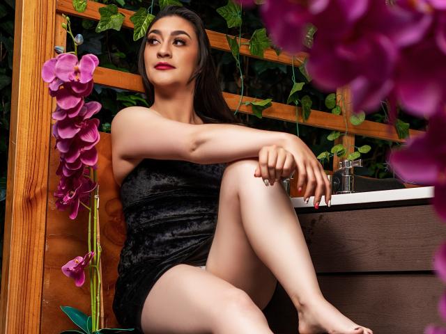 EmmaLovesX with pale skin, dark hair, red lipstick, black velvet dress, sitting with legs crossed, purple orchids in foreground, wooden fence, and green vines, looking up with a slight smile.