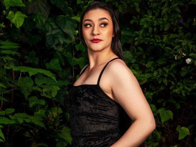EmmaLovesX with dark hair, red lipstick, and black lace dress, standing against green foliage, showing her tits and a confident ass-chieving smile. Her eyes are seductive and dark.