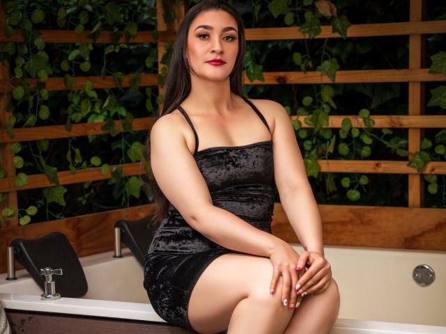 EmmaLovesX with pale skin, dark hair, and red lipstick, wearing a black velvet dress, sitting on a hot tub, hands on knees, showing her smooth thighs and small ass.