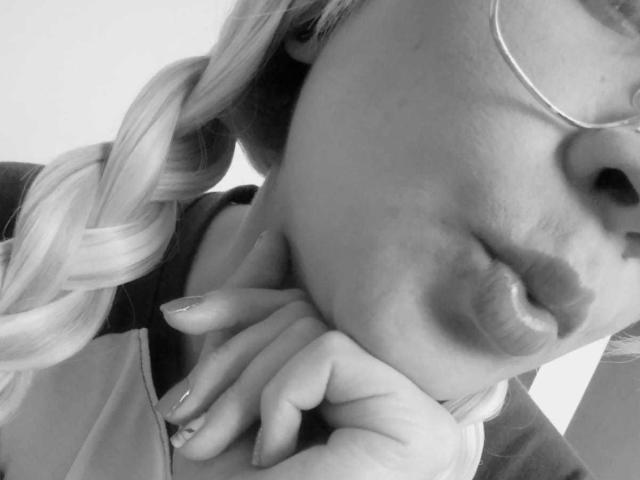 Selfie in bianco e nero di HornnyCoupleHot con labbra tumefatte, capelli biondi intrecciati, che indossa occhiali e un anello, con la mano vicino al mento, mostrando unghie curate..