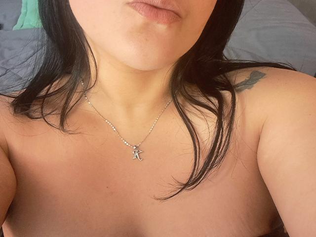 VivianaThompson has light skin, black hair, and is topless with a silver necklace featuring an "A" pendant. Her tits are partially visible, showing a small tattoo on her right shoulder.