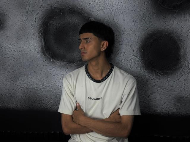 NathanMaxime, con capelli neri corti, pelle abbronzata e una maglietta bianca, braccia incrociate, che guarda di lato, con una parete grigia strutturata sullo sfondo, un piccolo piercing all'orecchio, espressione seria..
