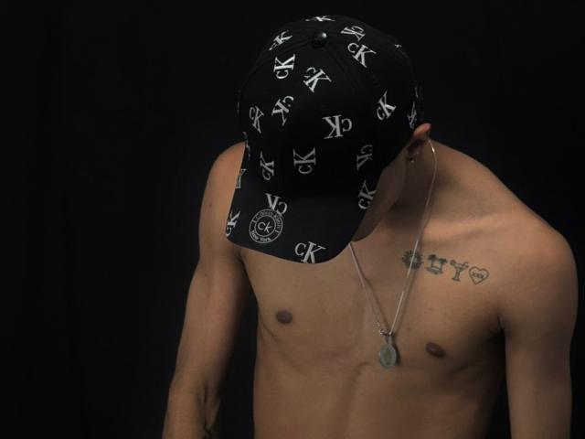 NathanMaxime, senza camicia, con un berretto nero, un portachinellino e tatuaggi, indossa un berretto con la scritta "RKR", con la testa bassa, mostrando un petto asciutto e muscoloso e piccoli capezzoli scuri..