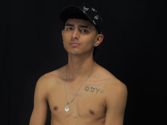NathanMaxime, ohne Hemd, mit einer schwarzen Mütze, brauner Haut, kleiner Brust, einer Halskette, einem Tattoo auf der rechten Brust und einem ernsten Gesichtsausdruck, vor einem dunklen Hintergrund..