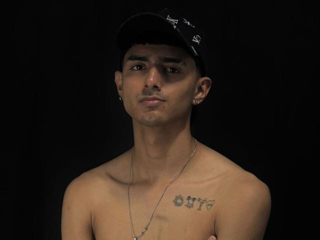 NathanMaxime, sem camisa e com um boné escuro, tem uma tatuagem com os números "187" e um sol no peito, usando um colar e brincos, com um fundo preto..