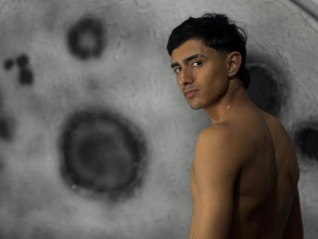 NathanMaxime, torse nu, avec des cheveux noirs courts, une peau brun moyen et un regard sérieux, se tient devant un arrière-plan gris flou évoquant la lune, montrant son dos musclé et un petit collier..