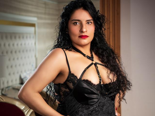 MoserratRouse with curly black hair, red lipstick, wearing black lace and satin lingerie with straps, standing in a room, looking directly at the camera, showing her tits.