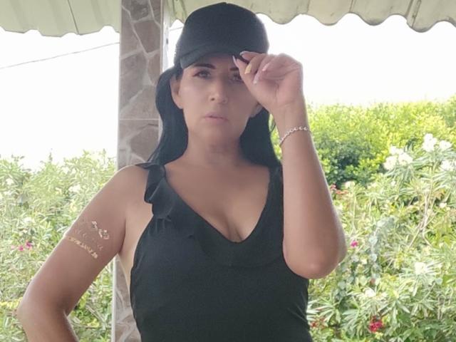 JackieMature with black hair, black tank top, ruffled sleeves, and bracelet, wearing a black cap, raising her cap slightly, showing her tits, standing in front of greenery.