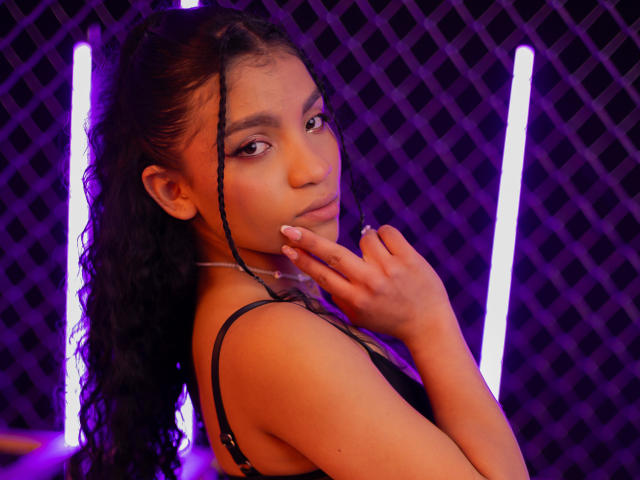 Eine Frau namens LightenStars mit lockigem, schwarzem Haar, brauner Haut und pinkem Lippenstift, der ihr Kinn berührt, trägt einen schwarzen BH und blickt in die Kamera. Im Hintergrund sind violette Neonlichter zu sehen..