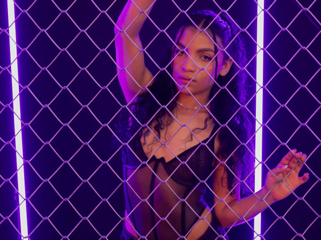LightenStars, con cabello negro rizado, vestida con una blusa de encaje negro transparente, con los brazos levantados y las manos sobre una cerca de alambre, luces de neón moradas, y una expresión segura y sensual..
