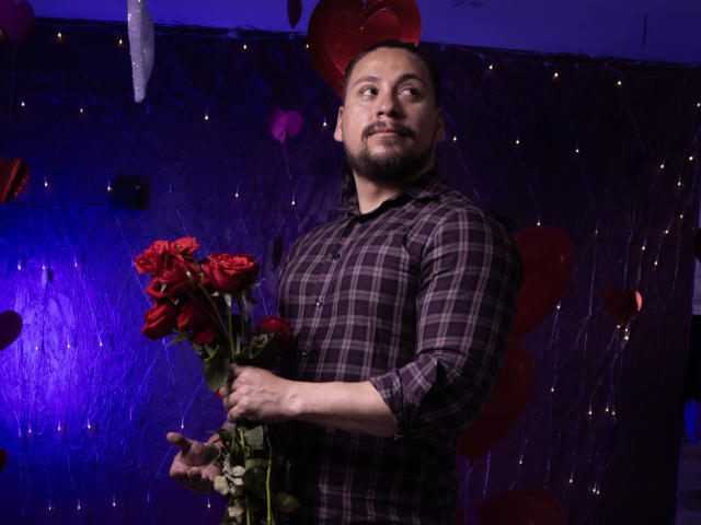 CameronDante with a beard, wearing a plaid shirt, holding a bouquet of red roses, standing in a room with purple lighting and red heart decorations.