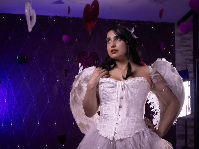 CameronDante, avec des ailes blanches en plumes, portant un corset blanc moulant qui expose sa poitrine généreuse, et une robe blanche et duveteuse, se tient debout avec assurance, une main sur sa hanche..