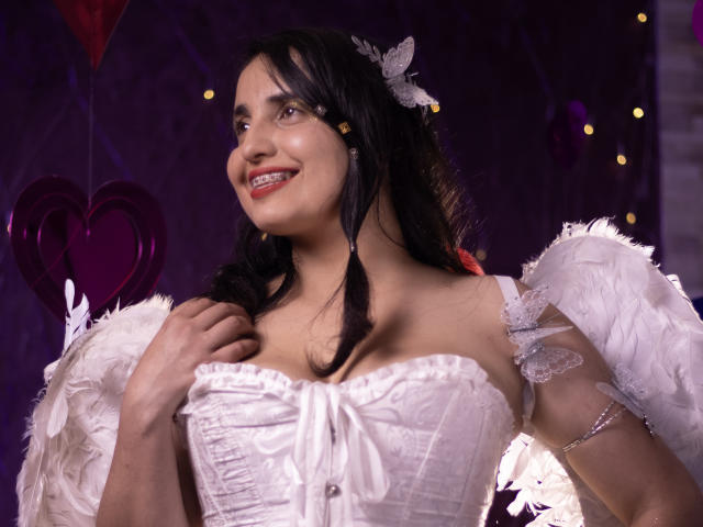 CameronDante with dark hair, white feathered wings, and a corset, smiling, holding her chest, white butterfly hairpiece, red lips, brown eyes, medium tits, purple background.
