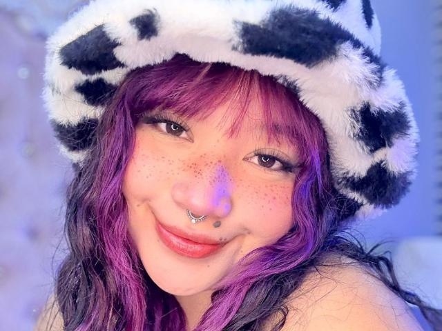 HonnyBlaice with purple and black hair, wearing a fuzzy black and white hat, smiling, has a nose ring, and a septum piercing, showing freckles and pink lipstick.