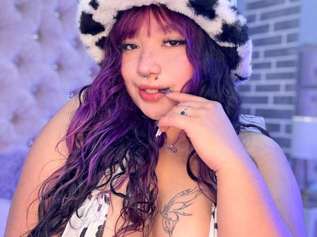 HonnyBlaice with purple and black hair, wearing a cow hat, has freckles, a nose ring, and a finger ring, showing a tattoo and her tits in a bedroom.
