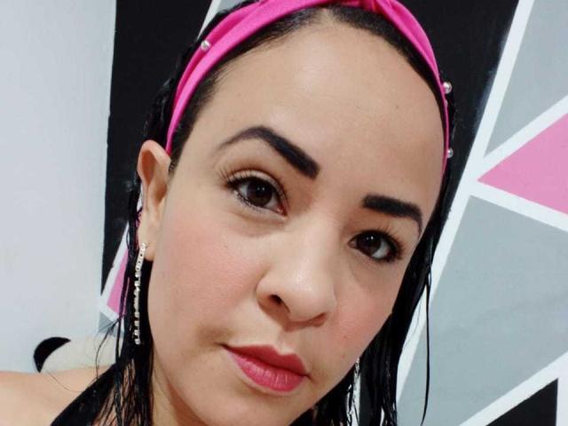 MimiP has tan skin, dark eyes, and pink lipstick. She's wearing a pink headband, black hair, and has earrings. Her eyebrows are thick, and she's looking at the camera.