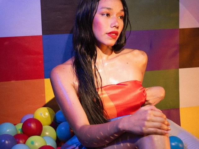 RoseSexAndFun with long black hair, red strapless top, sitting in a colorful balloon pile, arms crossed, showing her shiny tits and ass, looking sexy with red lipstick.