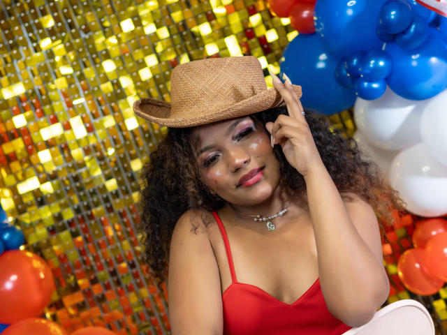 Eva Bennet, con cabello castaño rizado, vestido rojo y sombrero de paja, sonriendo, tocando el sombrero, labios rojos, maquillaje sutil, collar, pequeña tatuaje de estrella en el hombro, rodeada de globos azules y rojos..