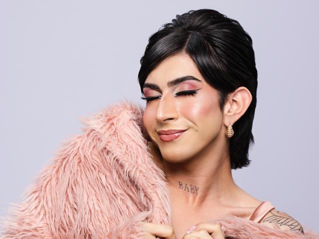 MayaPeitthon with short black hair, pink eyelids, and pink lipstick, hugging a fluffy pink pillow, smiling, wearing gold earrings, tattoo on neck, and pink top.