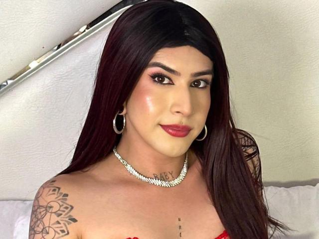 MayaPeitthon with long dark red hair, red lipstick, hoop earrings, diamond necklace, tattoo on shoulder, and subtle makeup, smiling at the camera, showing her bare tits.