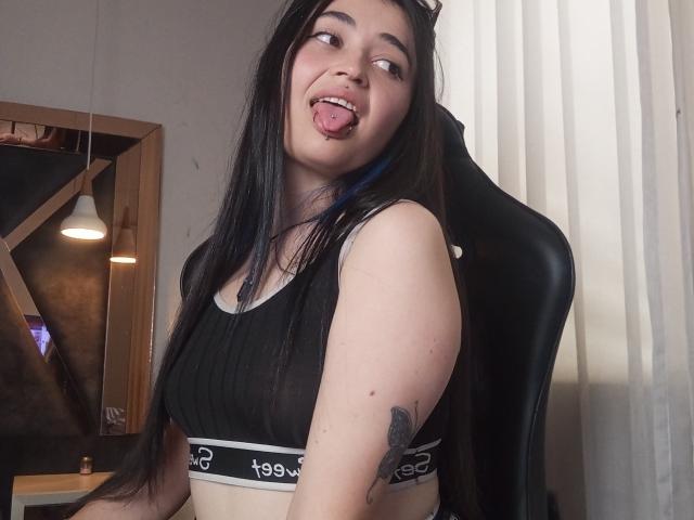 EvaMiller69 with long black hair, black sports bra, tongue out, tattoo on right arm, sitting in black chair, light skin, looking sideways, white curtain in background.