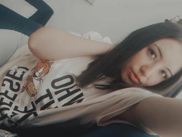 SusanMonroy with long dark hair, nose ring, and pouty lips, wearing a white tee, lying on a blue chair, arms resting on her head, soft light, slightly blurred.
