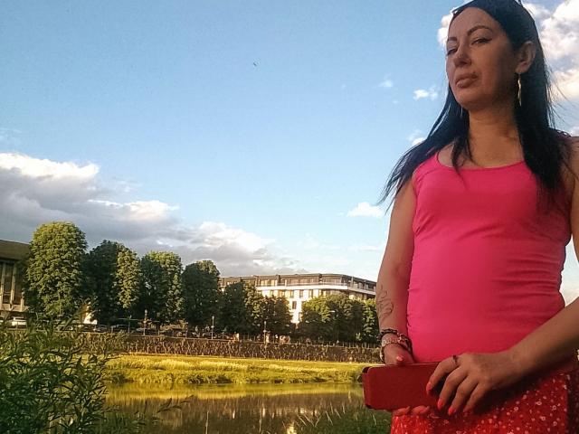 MirandaHott69 with long black hair, pink tank top, red skirt, holding red phone, standing by grassy pond, clear blue sky, white clouds, trees, and building in background.