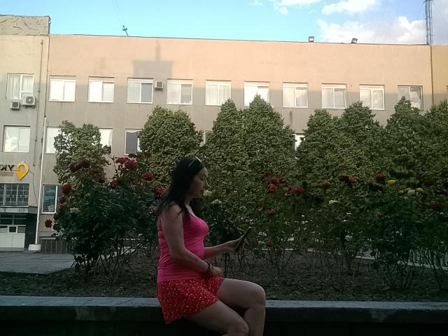 MirandaHott69 with tits in a pink tank top and red polka-dot ass in a skirt, sitting on a bench, texting on a phone, in front of a building.
