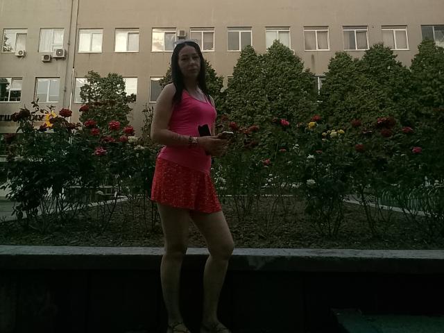 MirandaHott69 stands in front of a flower bed, wearing a pink tank top and red polka dot skirt, holding a black phone, with her tits slightly visible.