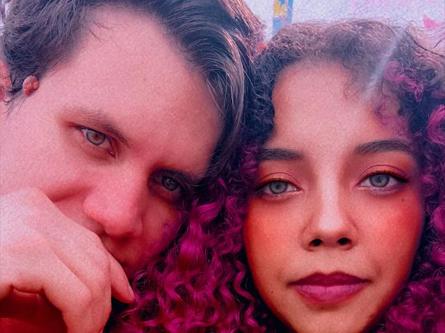 BethAndAnton's face with pink curly hair and blue eyes next to Anton's, who has brown hair and blue eyes. Anton's hand rests on BethAndAnton's cheek, both smiling.